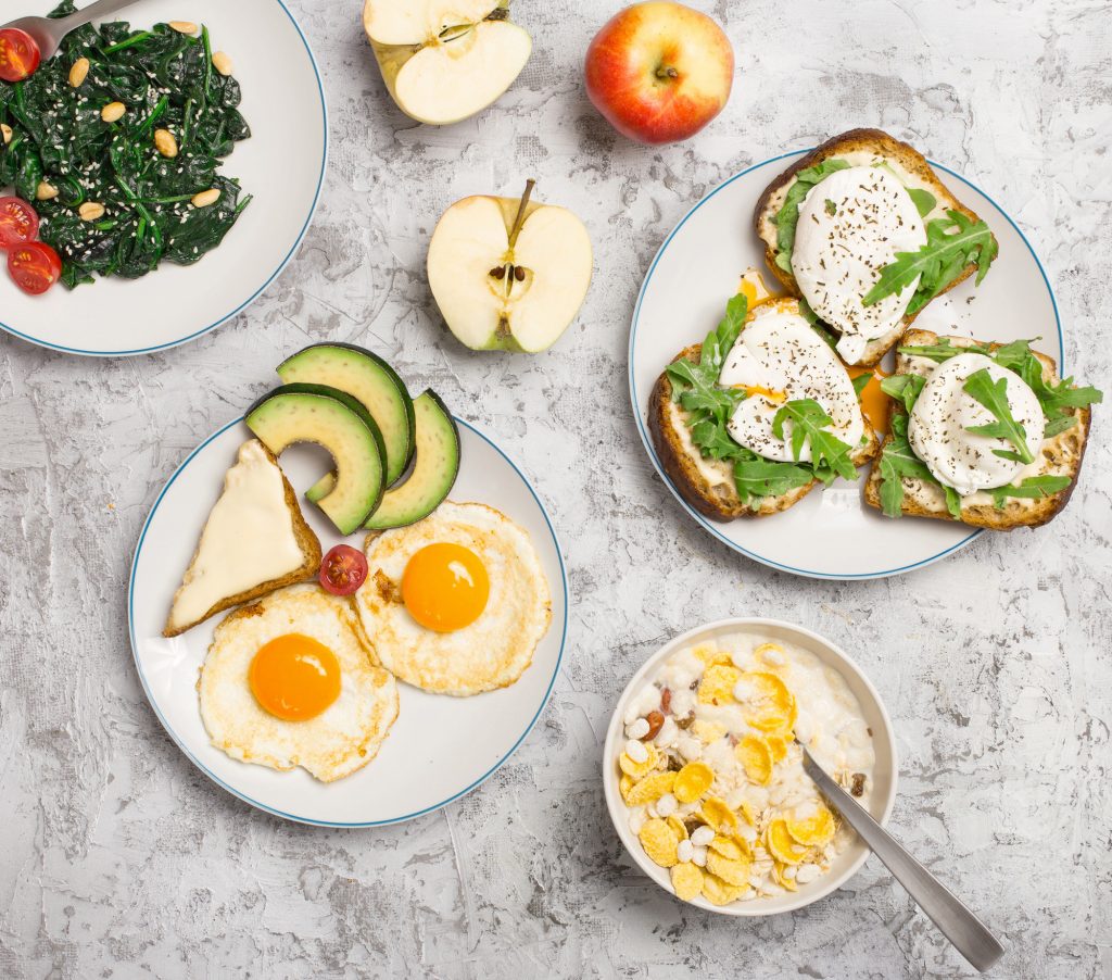 Helpful and tasty breakfast from different of dishes - fried egg, poached eggs, avocado, apple, spinach salad and muesli on light surface, top view