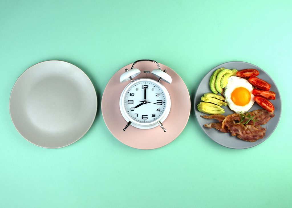 Intermittent fasting - three plates, one empty, one with a clock and one with a meal consisting of avocado, egg, tomatoes and bacon