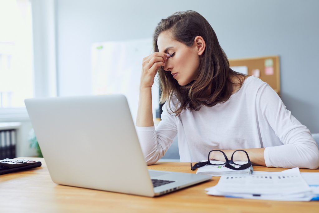 Young woman infront of a laptop looking stressed and holding her head in pain